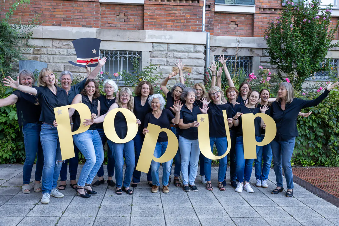 Der Frauenchor PopUp posiert mit den großen gelben PopUp-Buchstaben vor einer Backsteinwand.