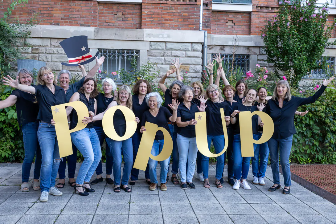 Der Frauenchor PopUp Schorndorf hält die großen gelben Holzbuchstaben P-O-P-U-P, im Hintergrund eine Backsteinwand.