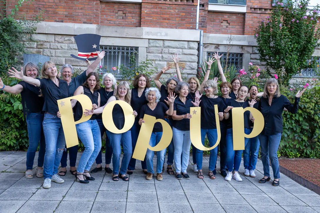 Der Frauenchor PopUp jubelt mit erhobenen Händen und den gelben PopUp-Buchstaben in die Kamera.